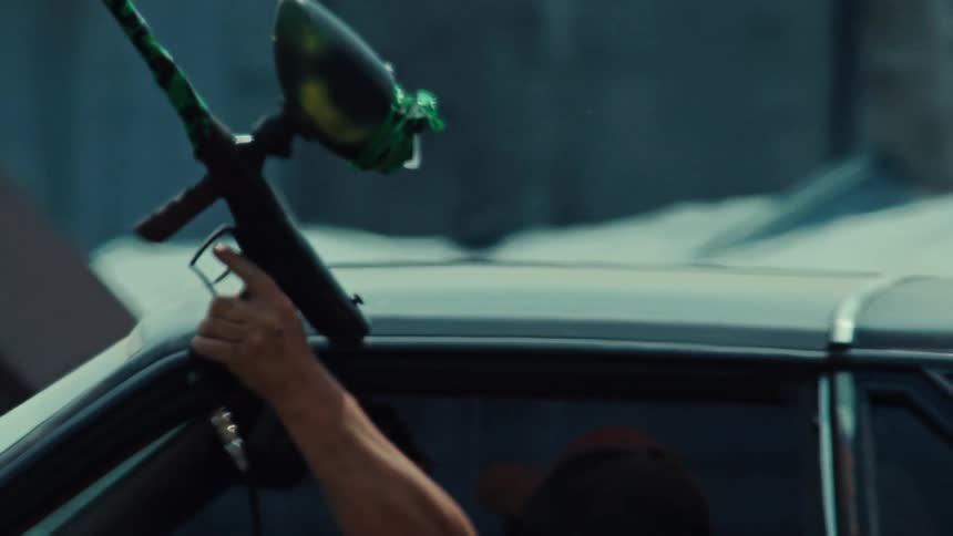 An arm holds a green and black paintball marker out of a car window before firing it in slow motion