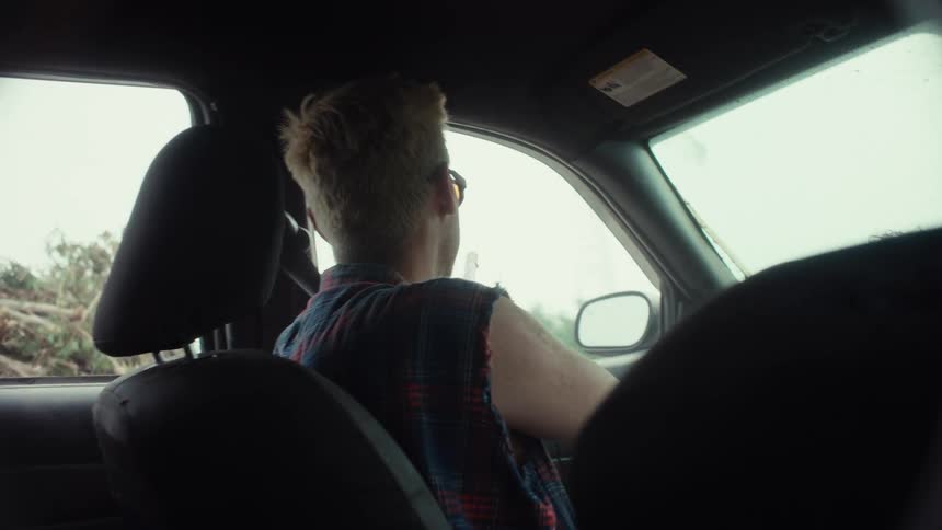 A punk rock-style man drives his car through a desolate landscape as seen from the backseat