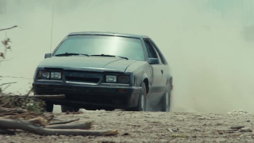 A man does a powerful burnout in a black vintage Mustang on a dirt road, creating a massive dust cloud in slow motion