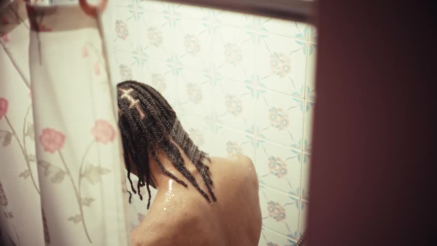 A young man with dreadlocks takes a relaxing and thoughtful shower, seen from behind a floral curtain