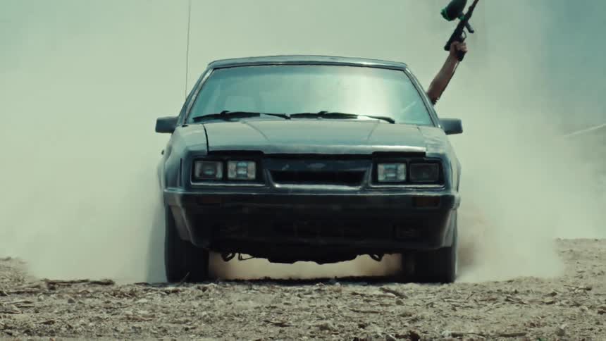A black Mustang does a smoky burnout as a person holds a paintball gun out the window