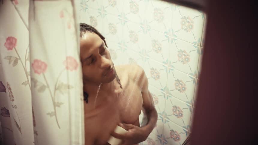 A man with dreadlocks washes his body with a bar of soap while taking a shower, viewed from behind a floral curtain