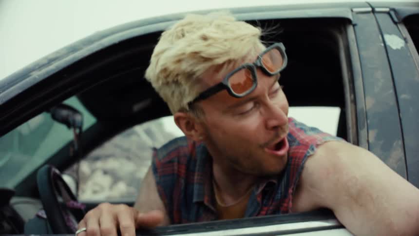 A man with bleached hair and sunglasses talks and smiles from his car in a junkyard before driving away in slow motion