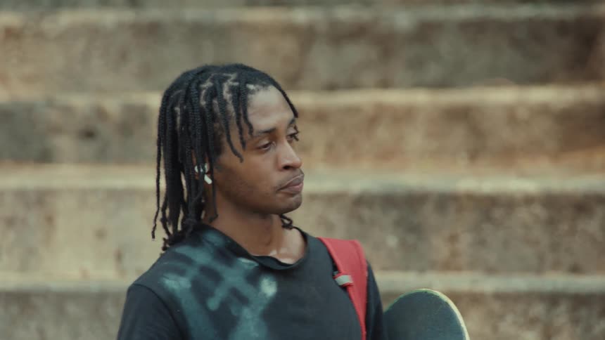 A slow-motion close-up pans up to a young Black man with a skateboard wiping his eyes while standing on concrete steps
