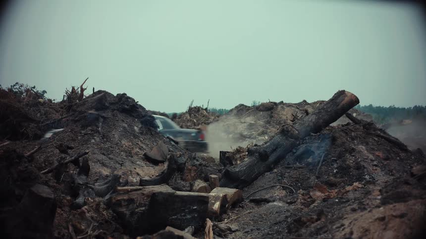 A beat-up car drives through a smoldering, desolate wasteland filled with burnt logs and debris