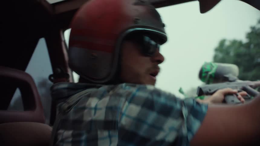 A man in a helmet and sunglasses drives a car while aiming a paintball gun out the window in a dynamic slow-motion shot