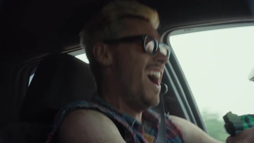 A wild, laughing man with dyed blond hair rides shotgun in a car while holding a paintball gun