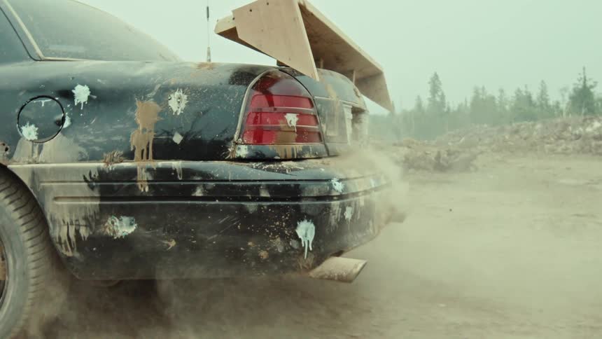 The exhaust pipe of a dirty black sedan with a crude wooden spoiler spews smoke and dust in a desolate landscape