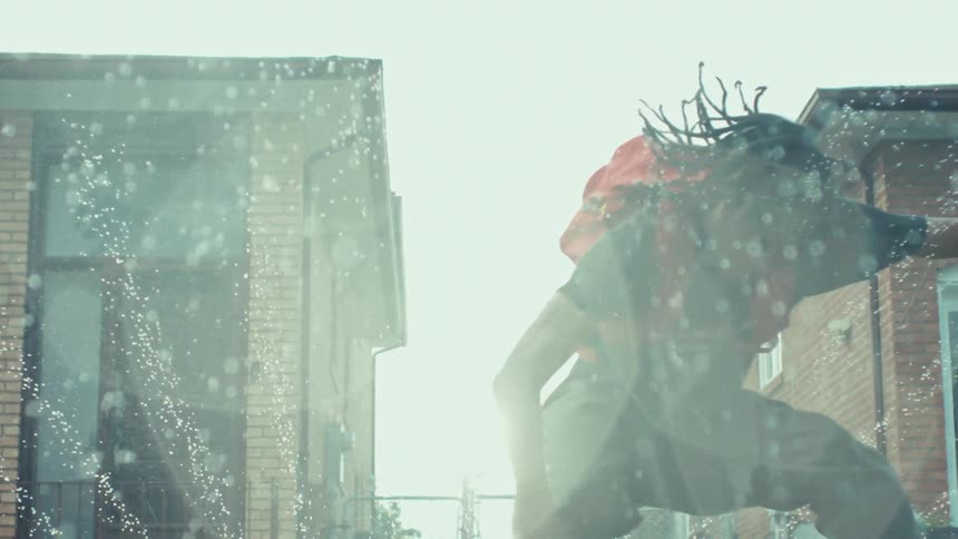 A young man with a red backpack playfully jumps through a sun-drenched sprinkler in a suburban backyard