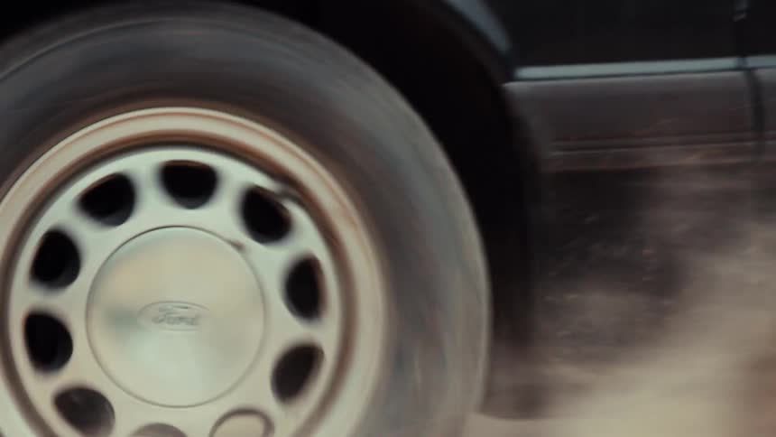 A close-up of a classic black muscle car doing a burnout on a dirt road, kicking up a large cloud of dust