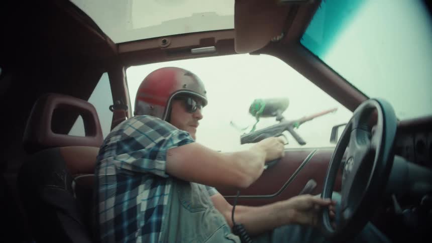 A man wearing a helmet and sunglasses drives a car erratically while holding a paintball gun in a post-apocalyptic setting