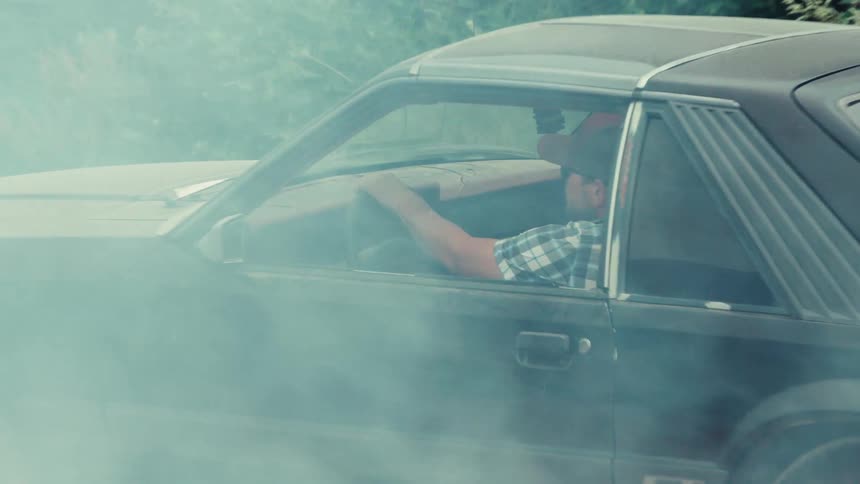Thick white smoke slowly clears to reveal a man in a classic black sports car after doing a burnout on a country road