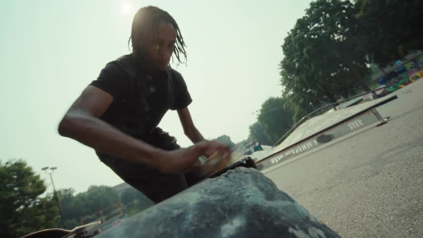 A focused skater with dreadlocks meticulously waxes a metal ledge at an outdoor skatepark in slow motion
