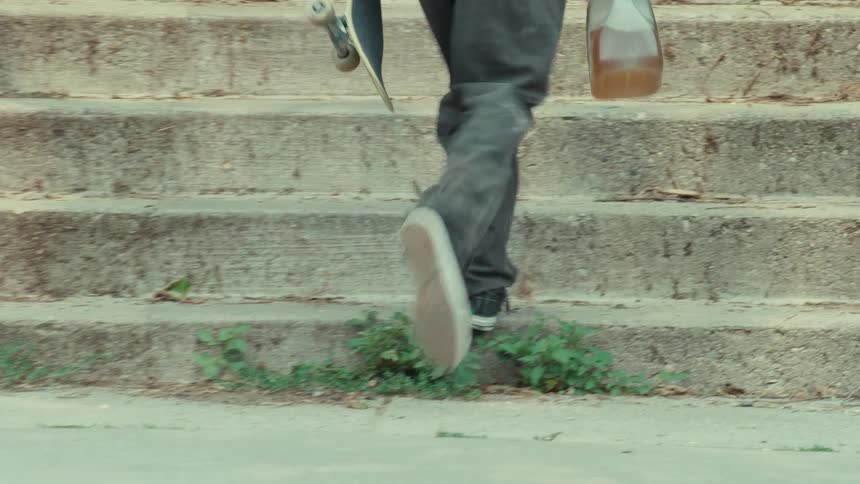 A tracking shot follows a person's feet as they skateboard on a sidewalk, before they pick up the board and walk up a set of concrete stairs