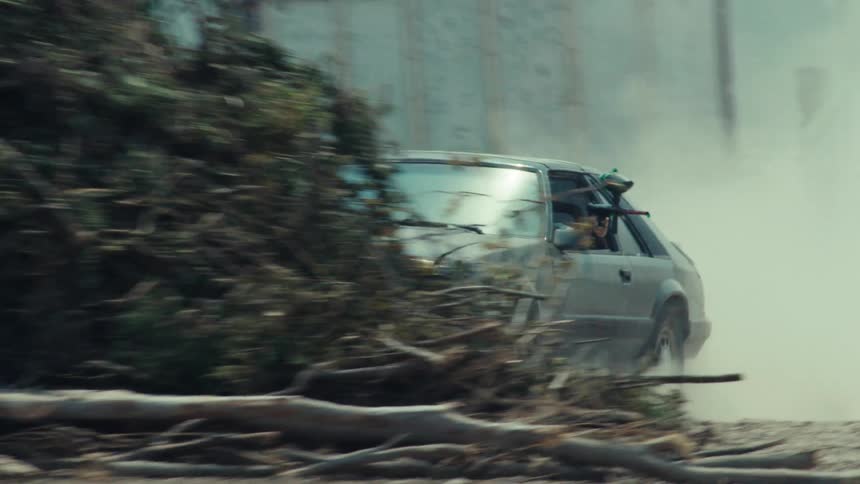 Two modified cars race in slow motion down a dusty rural road, with a passenger aiming a paintball gun from the window