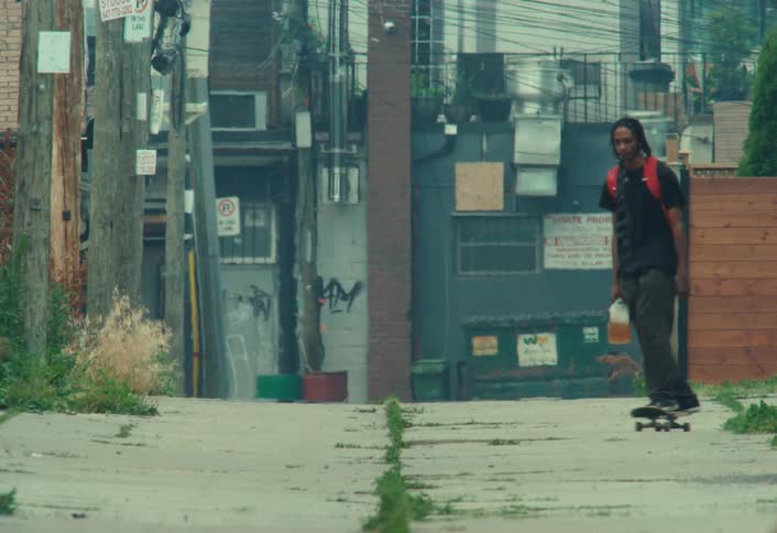 A young man casually skateboards down a gritty city alley while carrying a jug of iced tea and doing tricks