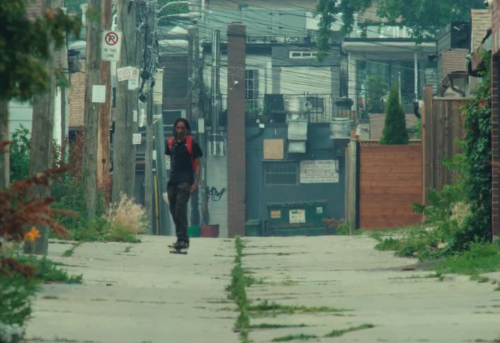 A cool young man with a red backpack and dreadlocks skateboards down a gritty urban alleyway toward the camera in slow motion