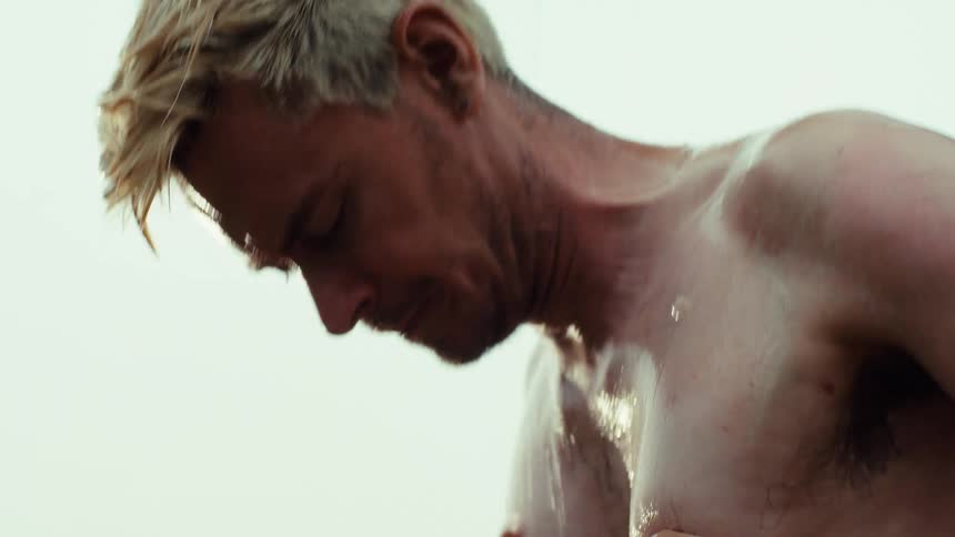 A shirtless, tattooed man with bleached blonde hair takes a vigorous outdoor shower in slow motion
