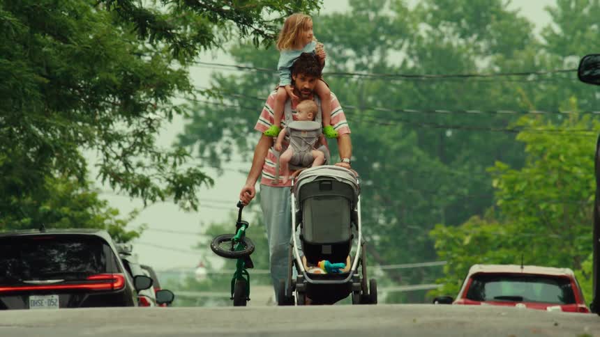 A busy father walks down a residential street with his toddler on his shoulders, a baby in a carrier, pushing a stroller, and carrying a small bike