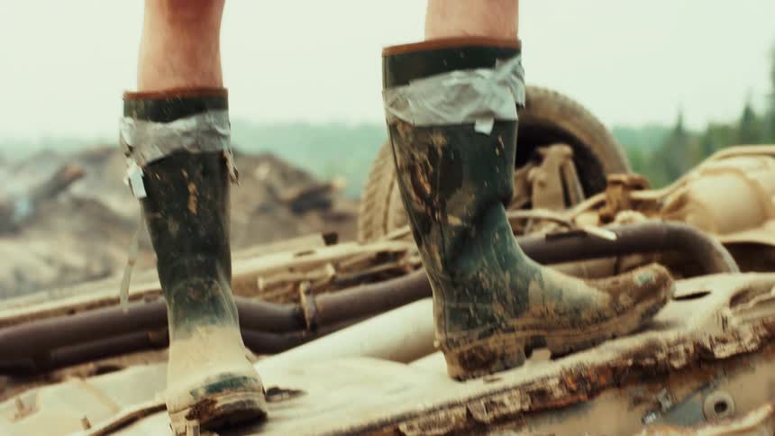 A young man in muddy rubber boots stands on a wrecked car in a junkyard and looks up at the sky