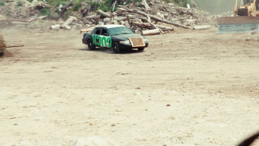 A classic black Mustang and a demolition derby race car drive through a dusty, industrial junkyard