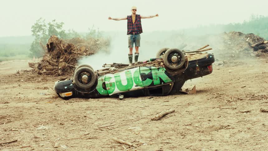 A rebellious man raises his arms in triumph while standing on a smoking, overturned car with graffiti in a desolate wasteland
