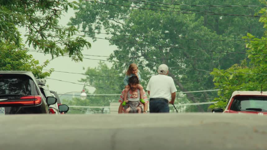 A father walks up a residential street carrying his toddler on his shoulders and his baby in a carrier while pushing a stroller