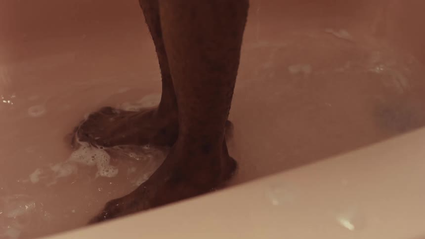 A person washes their feet in a soapy pink bathtub, dispensing soap from a bottle