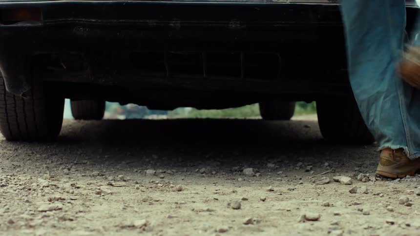 A person in dirty jeans and work boots stands behind an old car on a dirt road before walking out of frame