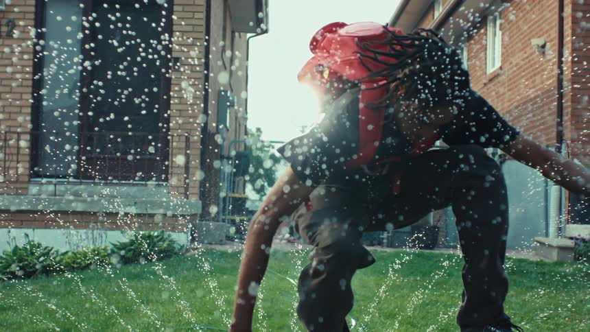 A young man with a red backpack playfully dances in a lawn sprinkler in a suburban backyard in slow motion