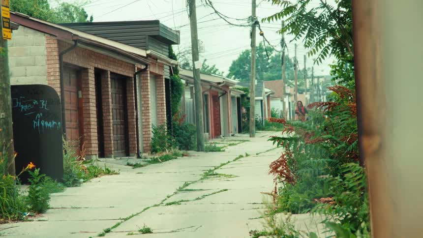 A lone skateboarder cruises down a long, overgrown urban alleyway lined with graffiti and residential garages