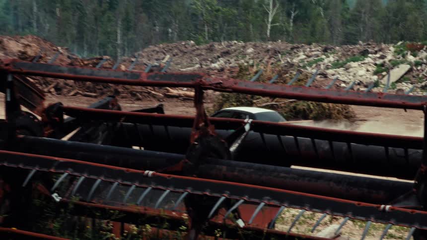 A car with 'GUCK' painted on its side speeds through a messy, rural lot filled with old farm equipment and debris