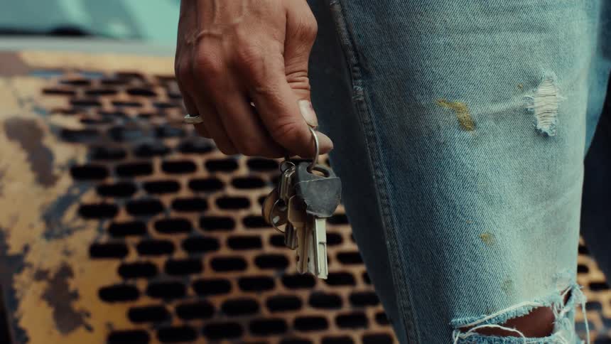 A close-up shot on a person's hand holding and jiggling a set of keys, next to their ripped and stained jeans