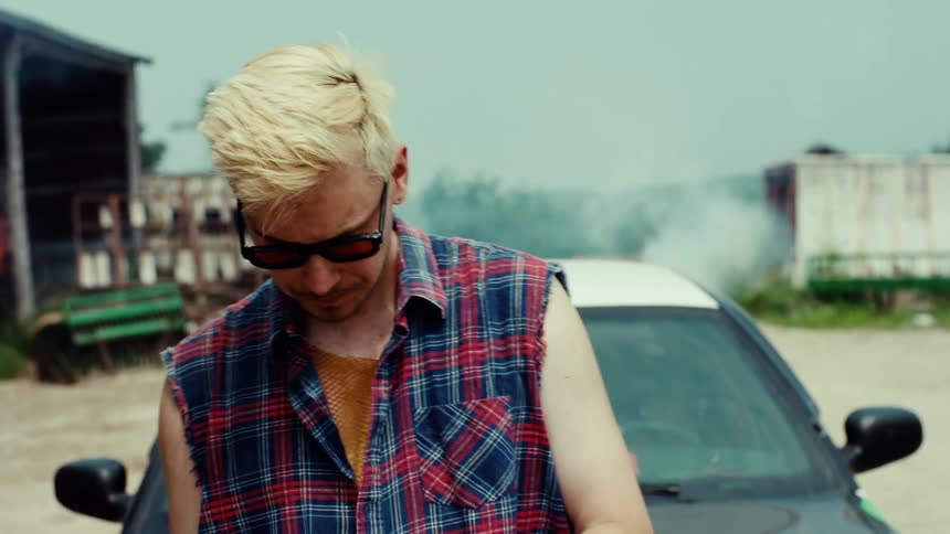 A stylish young man with bleached blonde hair and sunglasses stands confidently in front of a car in a rural, industrial setting