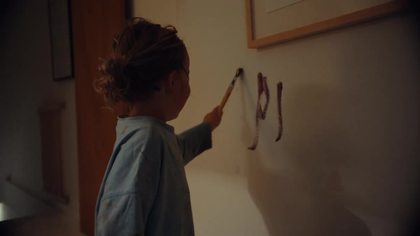 A cute and mischievous little girl with paint on her face and shirt joyfully paints on the wall inside her house
