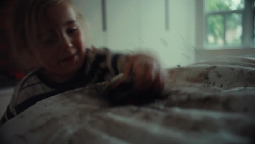 A young, muddy boy happily plays with his toy monster truck on white bedsheets covered in dirt