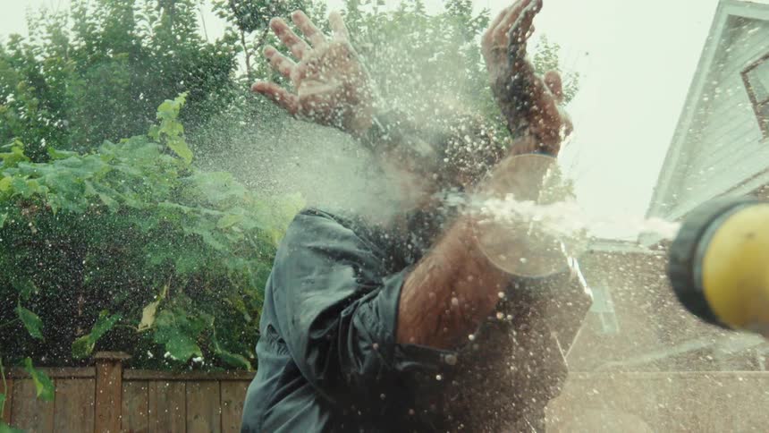 A man covered in mud shields his face while being sprayed with a garden hose in super slow motion