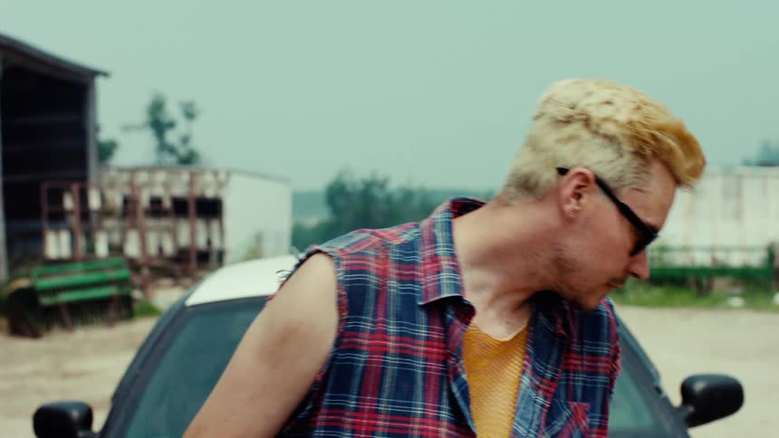 A rebellious-looking young man with bleached blond hair and sunglasses stares defiantly in a rural setting