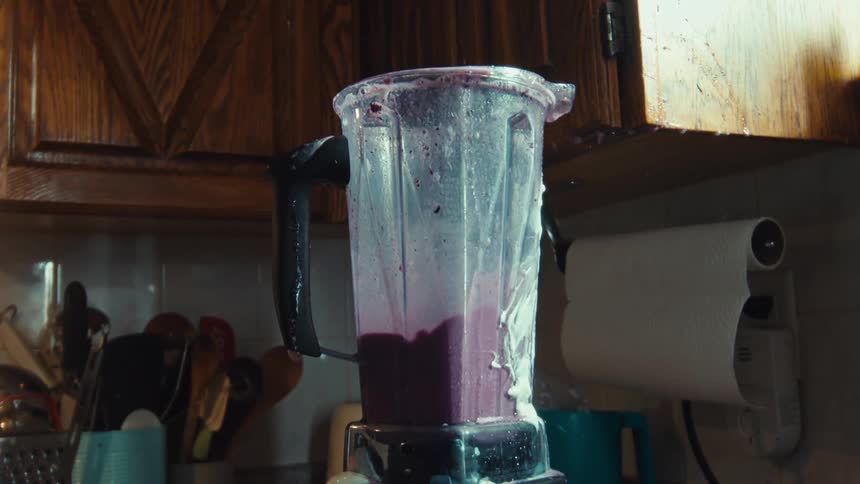 A messy blender churns a purple fruit and milk smoothie in slow motion in a home kitchen
