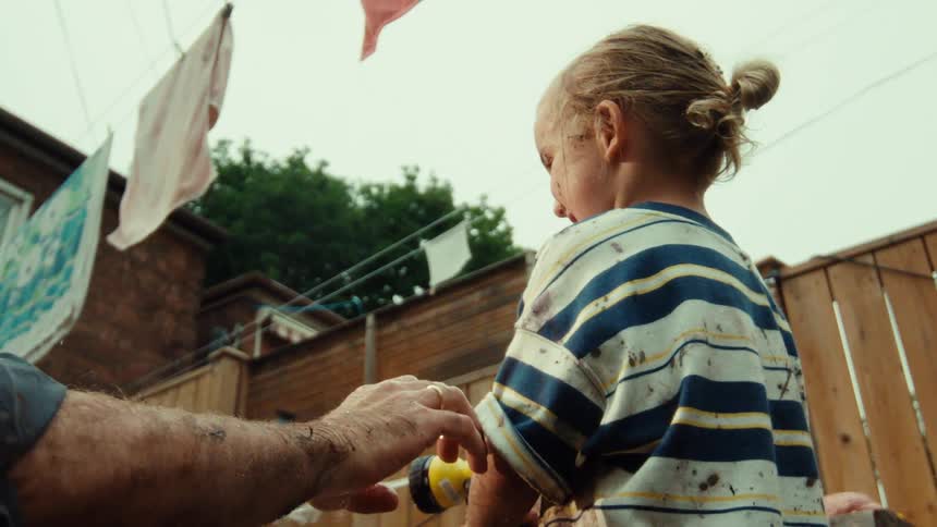 A father helps his muddy child spray a garden hose in their backyard in slow motion