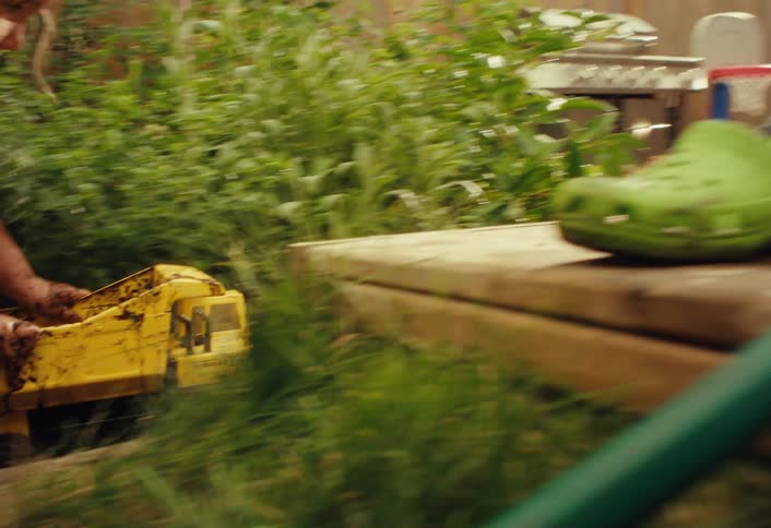 A happy young child covered in mud playfully pushes a yellow dump truck through the garden as their father watches on