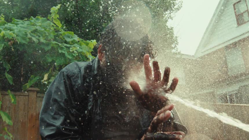 A man covered in mud shields his face while being sprayed with a hose in a backyard in dramatic slow motion