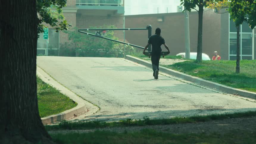 A lone skateboarder casually walks up a paved hill away from the camera in a quiet, green neighborhood
