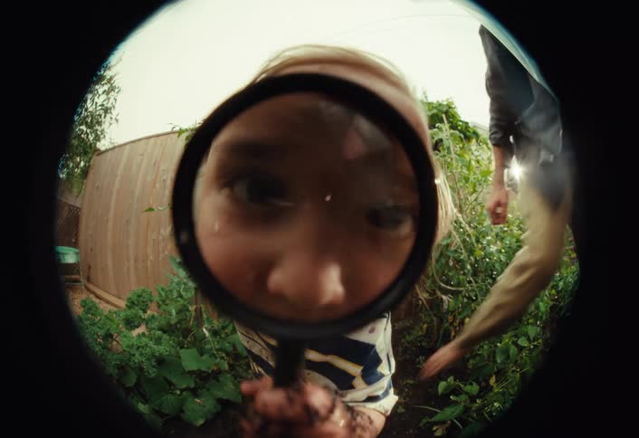 A curious toddler covered in mud holds a magnifying glass up to a fisheye lens while playing in the garden with their father