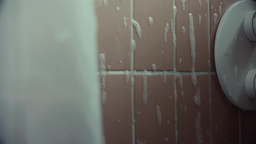 Close-up of soapy foam being sprayed onto a tiled shower wall and dripping down