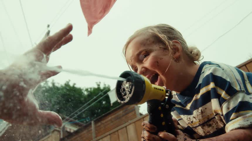 Low-angle slow-motion shot of a laughing, muddy girl having a water fight and spraying the camera lens in the backyard