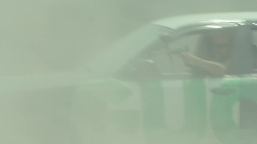 A modified race car with a paintball gunner drifts in slow motion, kicking up a massive cloud of dust on a dirt road