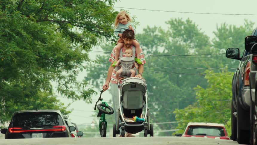 A multitasking dad walks down a suburban street, carrying his baby and toddler while pushing a stroller and holding a small bicycle