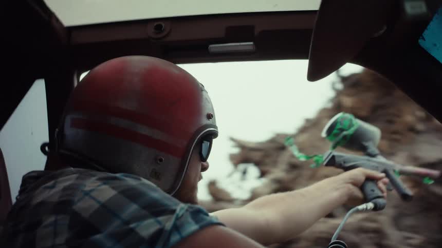 A man in a helmet and sunglasses drives a car with one hand and holds a paintball gun with the other during a slow-motion action sequence
