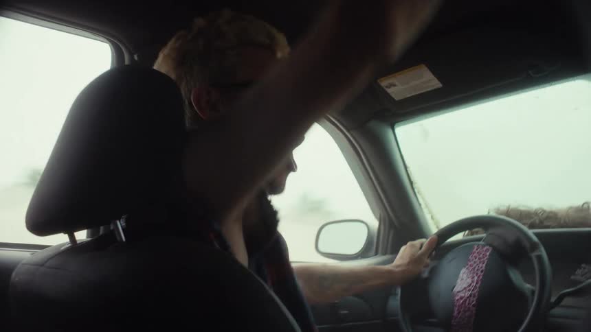 An energetic man with blonde hair and sunglasses yells and dances while driving his car through a severe storm